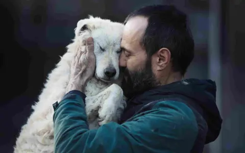 Un homme barbu enlace tendrement un chien blanc aux yeux fermés. Un moment de pur candilat chien.